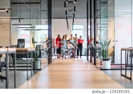 A diverse group of young business people walking a corridor in the glass-enclosed office of a modern startup, including a person in a wheelchair and a woman wearing a hijab 114165191