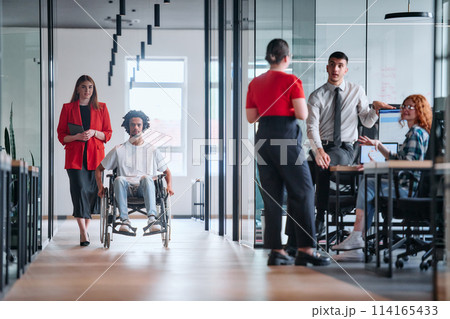 A business leader with her colleague, an African-American businessman who is a disabled person, pass by their colleagues who work in modern offices 114165433