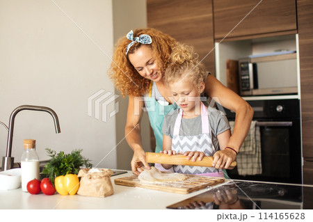 Beautiful young mom and her cute little daughter rolling out dough at home. 114165628