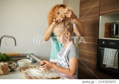 Little girl learn to make dough and her caring mother tying her hair up. Little girl learn to make dough and her caring mother tying her hair up. 114165655