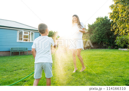 Joyful kid with mother playing with water sprinkler hose, splashing, laughing, having fun together in garden on sunset. Summer outdoor activity. Happy active childhood, family time. Selective focus 114166310