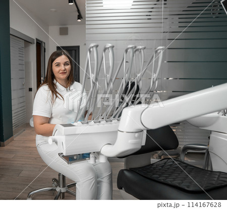A dentist sits near a dental chair, ready to provide assistance to future patients of a dental clinic. 114167628