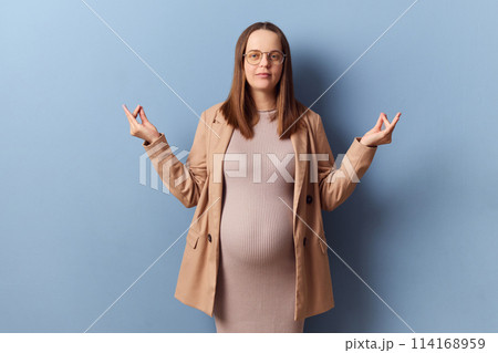Calm relaxed pregnant elegant woman in beautiful dress and jacket posing isolated over blue background standing with mudra gesture meditating trying to calm down 114168959