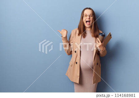 Excited pregnant woman in beautiful dress and jacket posing isolated over blue background showing advertisement area pointing at copy space for advertisement exclaiming happily 114168981