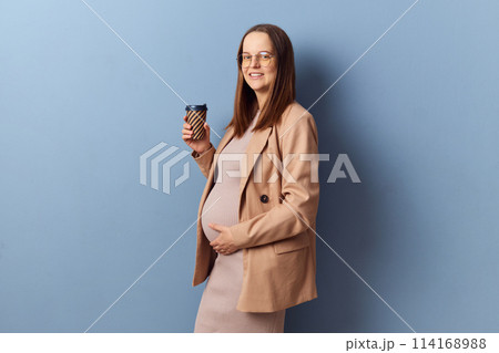 Smiling joyful pregnant woman in beautiful dress and jacket posing isolated over blue background enjoying hot takeaway coffee looking at camera drinking hot beverage 114168988