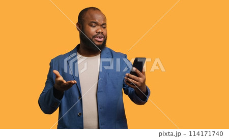 Upbeat man having friendly conversation with friends during teleconference meeting using smartphone, studio background. BIPOC person has fun catching up with mates during online videocall, camera A 114171740