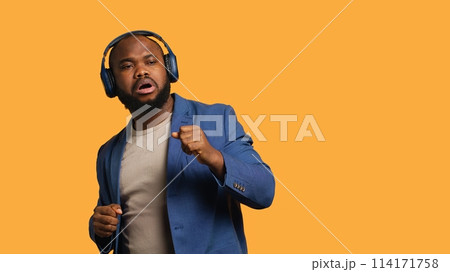 Carefree man listening energic music, raving on rhythm, enjoying free time. Euphoric BIPOC person wearing headphones, doing dance movements, isolated over studio background, camera A Carefree man listening energic music, raving on rhythm, enjoying free time. Euphoric BIPOC person wearing headphones, doing dance movements, isolated over studio background, camera A 114171758