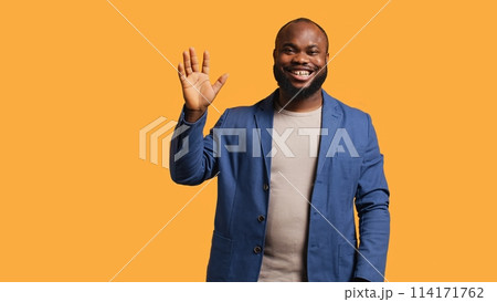 African american man cheerfully doing salutation hand gesture. Portrait of happy BIPOC person raising arm to greet someone, gesturing, isolated over yellow studio background, camera A 114171762