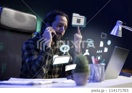 Smiling man talking on phone with coworker, working on laptop, accessing digital information concept. Employee using AR tech to interact with notebook, chatting over telephone call 114171765