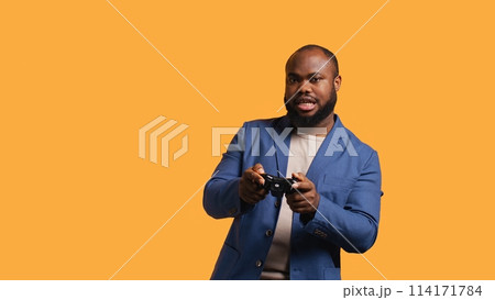 Upset man showing thumbs down sign gesturing holding controller, disapproving of videogames. Displeased BIPOC person against gaming, doing rejection hand gesture, studio background, camera B 114171784