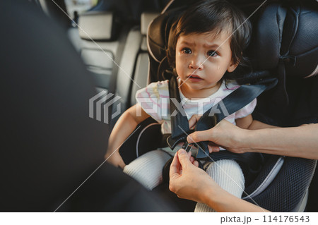 mother is fastening safety belt to crying toddler girl in car seat, safety baby chair travelling 114176543