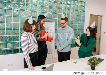 Friendly team of multiethnic business people congratulate African-American female colleague and give gift with surprise. Cheerful birthday woman surprised to open box with gift by around coworkers. 114180593