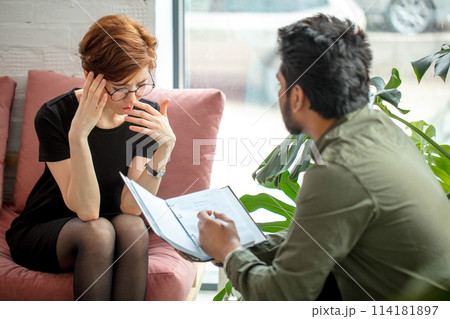 woman is sitting on couch and talking to male indian psychologist while doctor making notes woman is sitting on couch and talking to male indian psychologist while doctor making notes 114181897