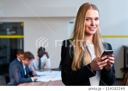 Confident blonde businesswoman uses smartphone in modern office, colleagues work at table behind. Pro female in smart casual attire interacts with mobile app for corporate tasks. 114182240