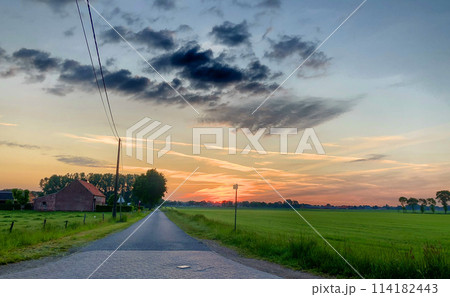 Dramatic Sunrise Over Rural Road with Farmhouse and Lush Fields 114182443