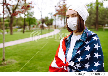Girl in protective sterile medical mask and with american flag in hand. Quarantine city. Covid -19. Girl in protective sterile medical mask and with american flag in hand. Quarantine city. Covid -19. 114188334