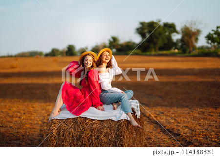 Two smiling young woman resting near haystack. Fashion concept. Nature, vacation, relax and lifestyle. 114188381