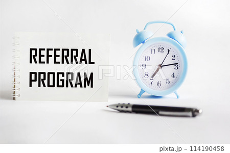 Notepad with the inscription REFERRAL PROGRAM on a white background with a clock and a pen. t. 114190458