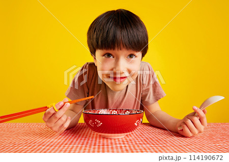 Cheerful young boy using chopsticks and spoon, sitting at table and eating ramen, noodle soup against yellow background 114190672