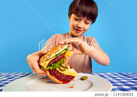 Little Korean 7 ears old boy eating giant juicy burger with fresh vegetables and cheese against blue background. Fast food dinner Little Korean 7 ears old boy eating giant juicy burger with fresh vegetables and cheese against blue background. Fast food dinner 114190691
