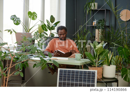 Portrait of adult African American man reading notes in planer while sitting at workplace with lush green plants copy space 114190866
