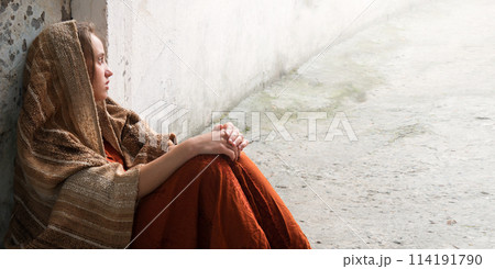 A girl in poor clothes sits on the ground 114191790
