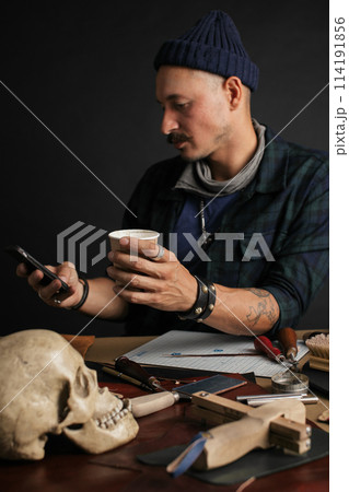 Leather Craftsman using smart phone and holding cup of coffee indoors Leather Craftsman using smart phone and holding cup of coffee indoors 114191856