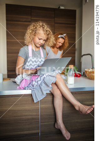Little girl and her daughter baking cookies at home, reading recipe on laptop. Little girl and her daughter baking cookies at home, reading recipe on laptop. 114194776