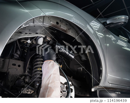 An auto mechanic applies anti-corrosion mastic to the underbody of a car. 114196439