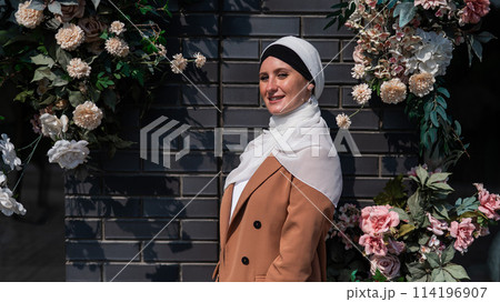 Portrait of a young caucasian woman dressed in a hijab near a wall with flowers. 114196907