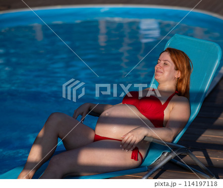 Red-haired pregnant woman in a red bikini sunbathes lying on a sun lounger next to the pool. Red-haired pregnant woman in a red bikini sunbathes lying on a sun lounger next to the pool. 114197118
