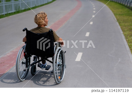 Rear view of an elderly woman in a wheelchair riding on a bike path. Rear view of an elderly woman in a wheelchair riding on a bike path. 114197119