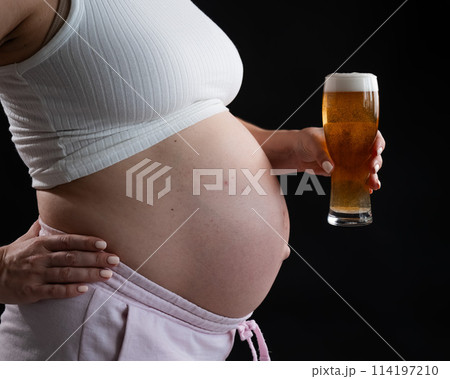 Close-up of the belly of a pregnant woman holding a glass of beer on a black background. Skin rash.  114197210