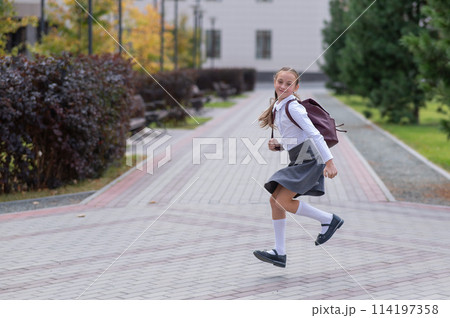 Happy caucasian girl in uniform and with a backpack jumping outdoors after school. 114197358
