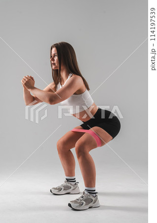 Caucasian woman doing squats with fitness band on white background. Vertical photo. Caucasian woman doing squats with fitness band on white background. Vertical photo. 114197539