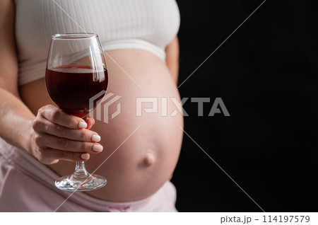 Close-up of the belly of a pregnant woman holding a glass of red wine on a black background.  114197579