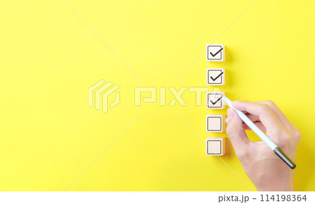 Checklist concept. Person using a black ink pen to correct sign and tick check mark on wooden block with copy space for the checklist on yellow background. Business survey questionnaire, exam process, Checklist concept. Person using a black ink pen to correct sign and tick check mark on wooden block with copy space for the checklist on yellow background. Business survey questionnaire, exam process, 114198364