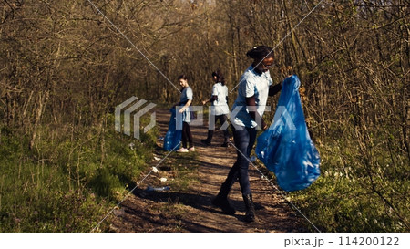 Group of diverse activists picking up the trash and plastic waste, collecting and recycling rubbish in the woods. People doing voluntary work to clean the natural habitat. Camera B. 114200122