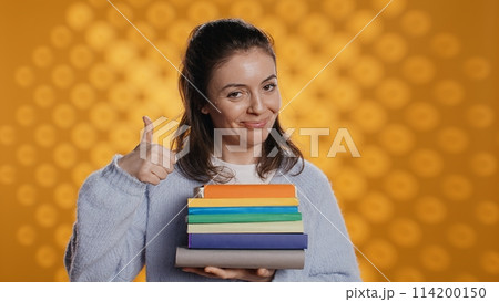 Portrait of happy woman with stack of books in hands showing thumbs up, studio background. Joyous bookworm holding pile of novels, feeling upbeat, doing positive hand gesturing, camera B Portrait of happy woman with stack of books in hands showing thumbs up, studio background. Joyous bookworm holding pile of novels, feeling upbeat, doing positive hand gesturing, camera B 114200150