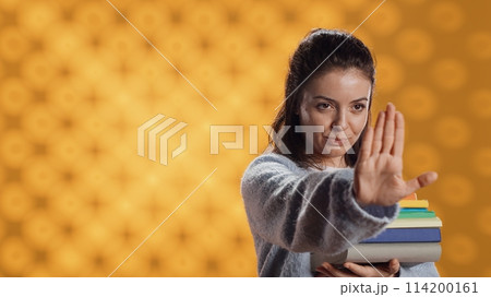 Portrait of stern woman holding stack of books doing stop sign gesturing, studio background. Student with pile of textbooks in arms used for academic learning doing halt hand gesture, camera A 114200161