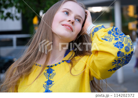 young beautiful woman, young girl in big city in yellow embroidered shirt, embroidered blouse, long brown hair, smile, feather earrings, yellow and blue colors in clothes, Ukrainian national costume 114202372