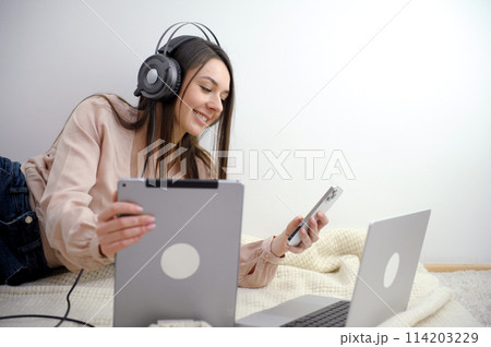 Smiling female student wears wireless headphones to study online with a Skype teacher, happy young woman learns a language, listen to a lecture watch a webinar, write notes, look at a laptop 114203229