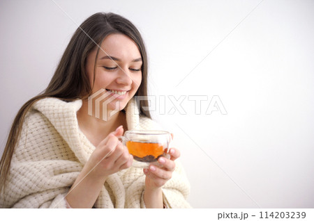 Happy young woman drinking a cup of tea in an autumn morning. Dreaming girl sitting in living room with cup of hot coffee enjoying under blanket with closed eyes. Pretty woman wearing sweater at home. 114203239