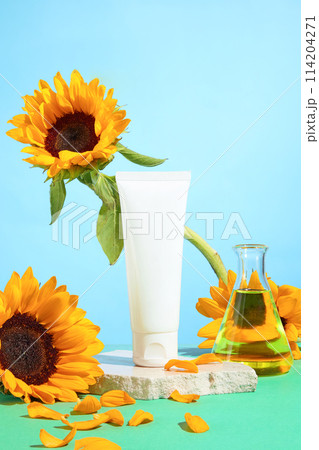 Blank label tube placed on white plaster podium, decorated by sunflower and lab glassware over light blue background. Space for advertising, front view 114204271