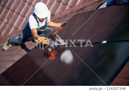 Worker building photovoltaic solar panel system on rooftop of house. Close up of man engineer in helmets and gloves installing solar module with help of hex key outdoors. Renewable energy. Worker building photovoltaic solar panel system on rooftop of house. Close up of man engineer in helmets and gloves installing solar module with help of hex key outdoors. Renewable energy. 114207346