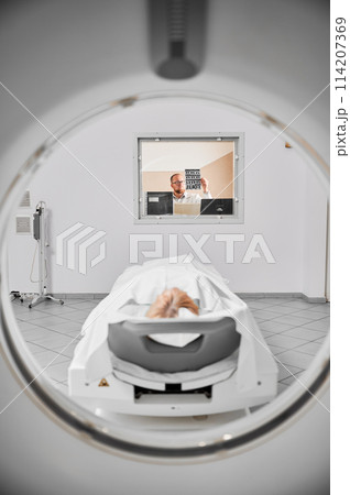 Medical computed tomography or MRI scanner. Doctor examining MRI results, female patient lying on couch. Doctor wearing glasses, holding MRI results. Concept of modern diagnostics. 114207369