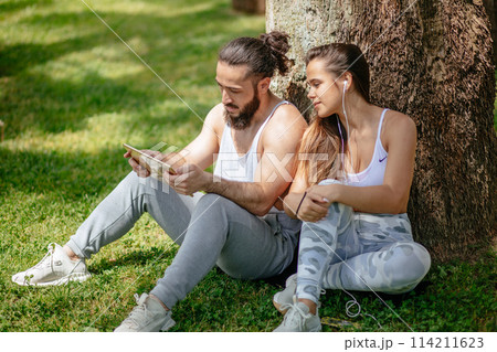 Man and woman relaxing with digital tablet in park after sports training 114211623