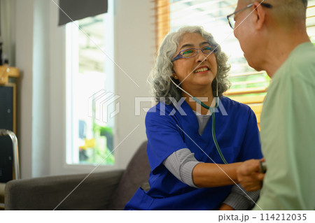 Smiling female doctor using stethoscope examining senior male during home care visit. Elderly healthcare concept 114212035