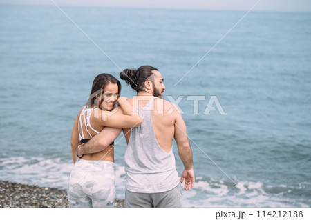 Young fitness couple embracing standing on beach, looking at horizon 114212188