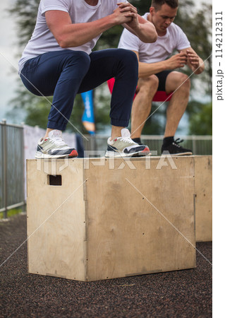 Two male athletic friends doing box jump outdoor on top of the mountain. Two male athletic friends doing box jump outdoor on top of the mountain. 114212311
